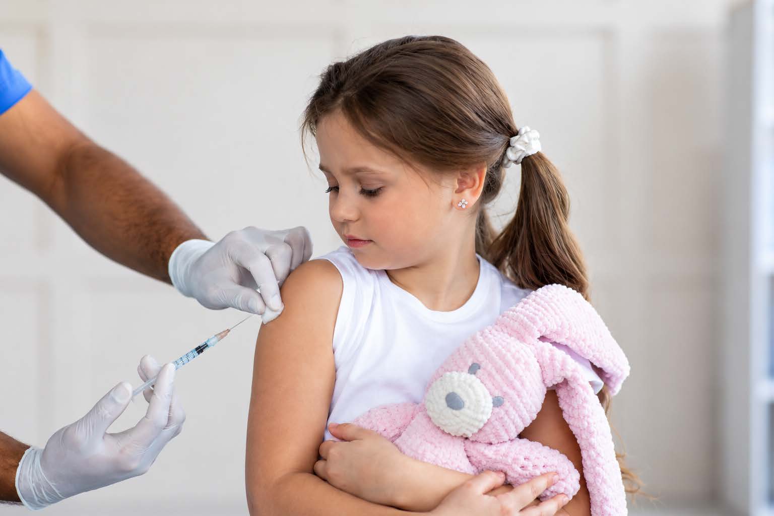 A young girl holding a stuffed rabbit receives a vaccine injection in her upper arm.